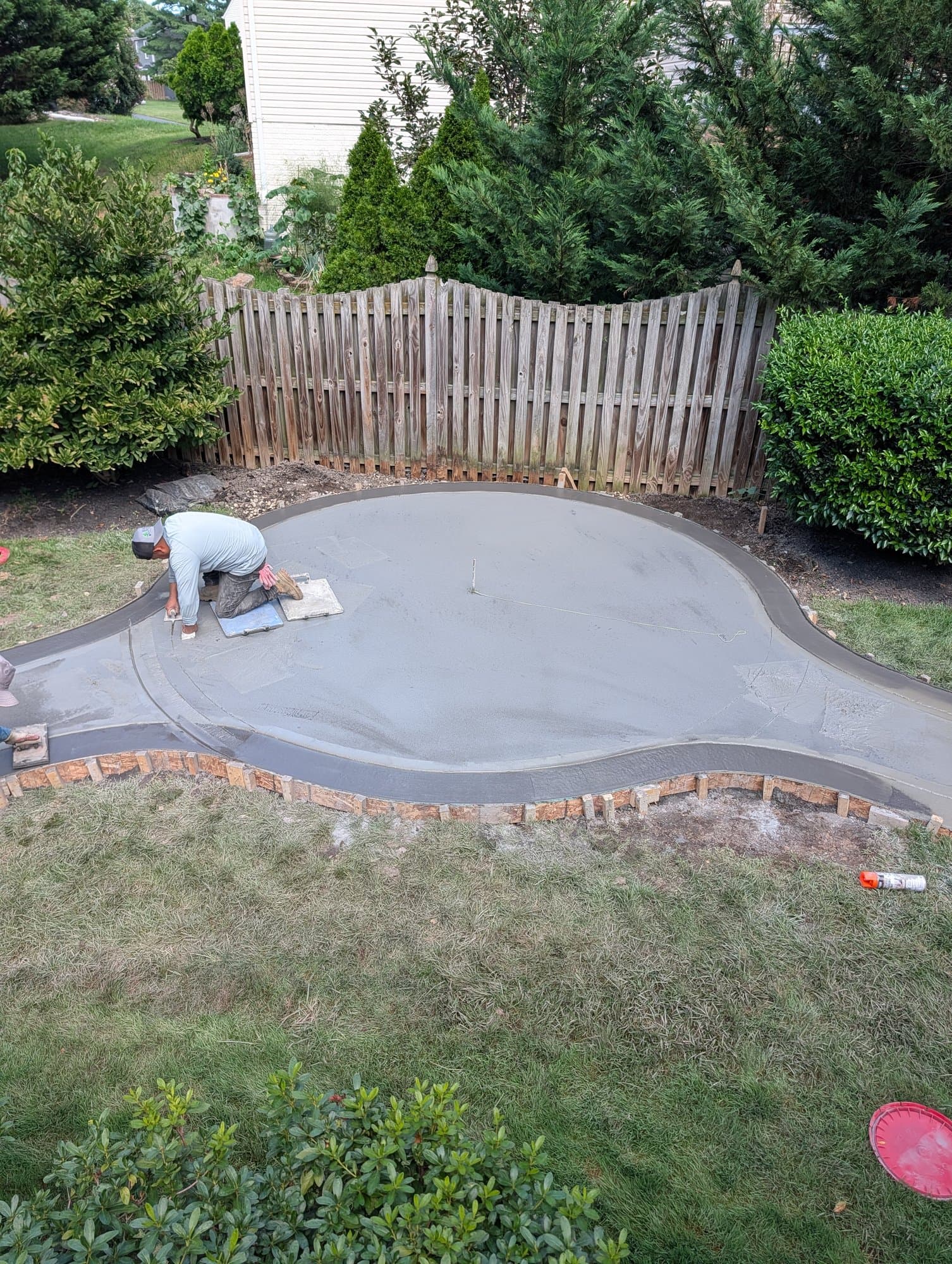 Aerial view of a large curved stamped concrete patio being hand-finished, with wood forms still in place along one edge, next to a wood privacy fence