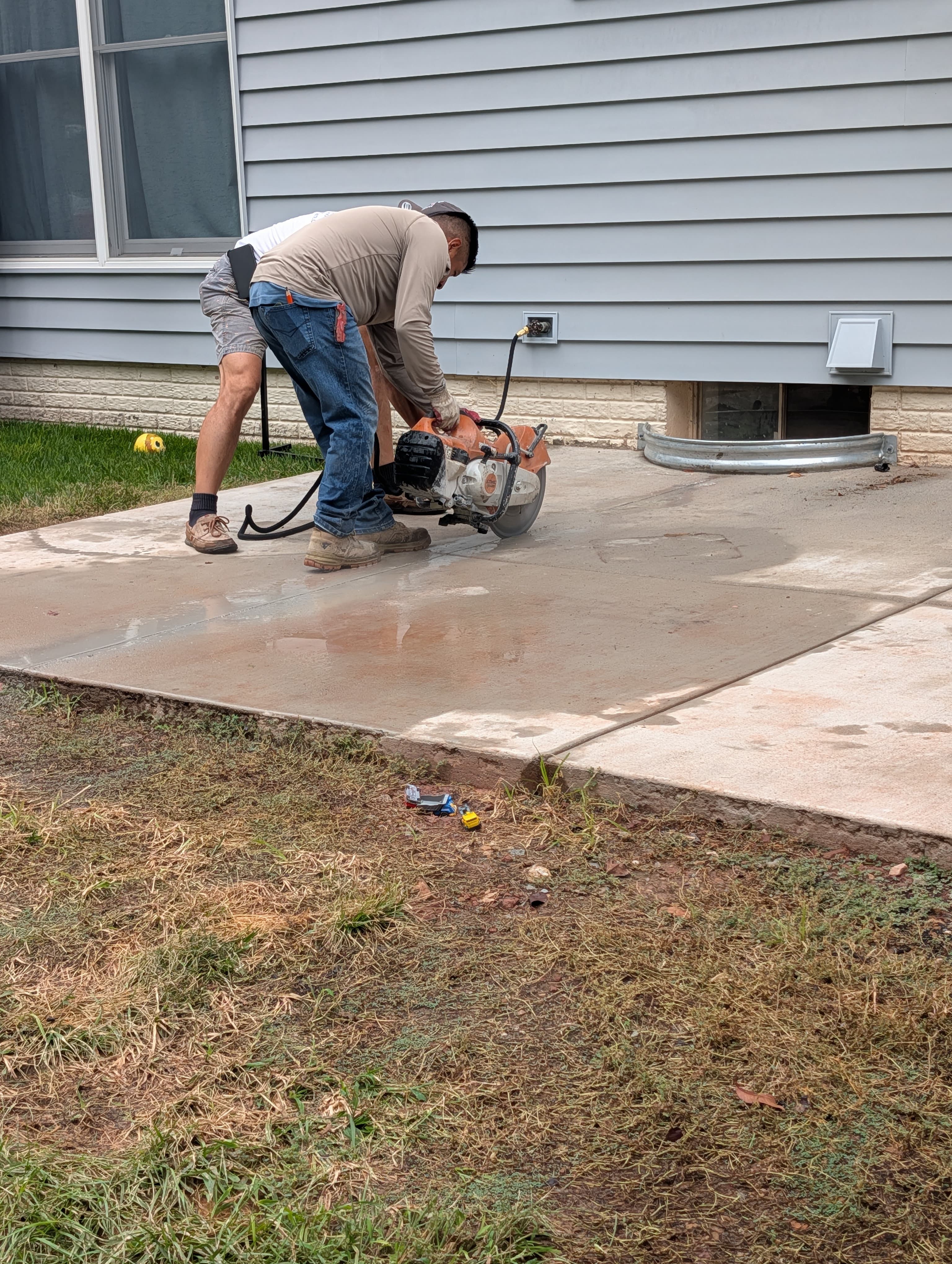 Worker using a gas-powered concrete saw to cut an existing slab next to a basement window well, with water for dust control